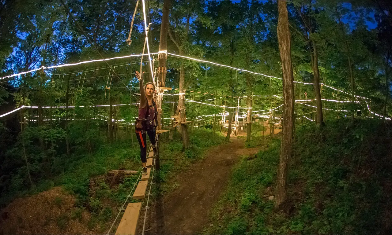 Woman participating in a night climb at TreEscape Aerial Adventure Park, wearing safety gear and navigating a lit obstacle course under the stars.