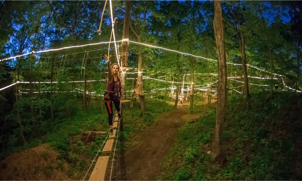 Woman participating in a night climb at TreEscape Aerial Adventure Park, wearing safety gear and navigating a lit obstacle course under the stars.
