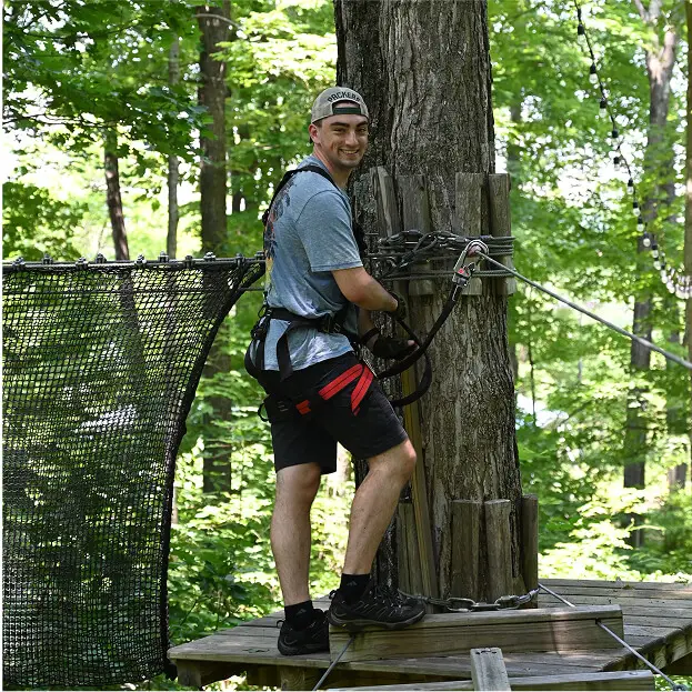 Young man smiling at TreEscape Aerial Adventure Park in Vernon, NJ, enjoying the outdoor adventure atmosphere surrounded by trees and activities.