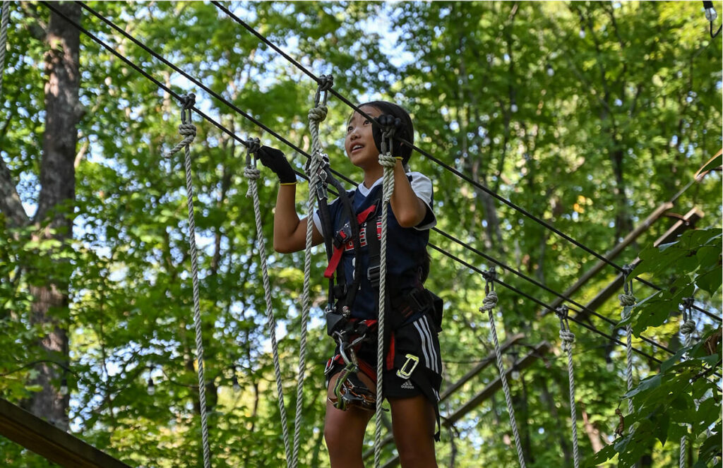 A girl, fully geared up, tackling the aerial ropes course at TreEscape Park in Vernon, NJ, relishing the excitement of the challenge high in the trees.
