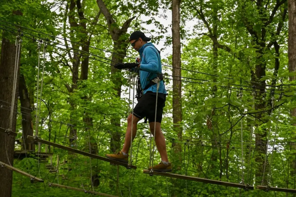 A man wearing sunglasses navigating a ropes course at TreEscape Aerial Adventure Park in Vernon, NJ, surrounded by towering trees and suspended obstacles.