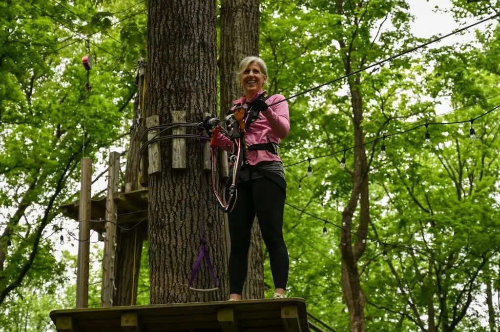 A woman preparing to start a ropes course at TreEscape Aerial Adventure Park in Vernon, NJ, adjusting her harness and getting ready for the challenge ahead, surrounded by towering trees, as a concept of ropes course near me.