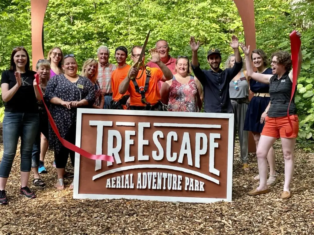 A group of people gathered around the TreEscape Aerial Adventure Park banner, displaying the park’s logo, excited and ready to begin their ropes course adventure, surrounded by towering trees and outdoor obstacles.