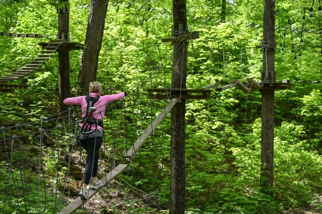 A woman photographed from behind navigating a ropes course at TreEscape Aerial Adventure Park in Vernon, NJ, carefully stepping across suspended obstacles surrounded by lush trees.