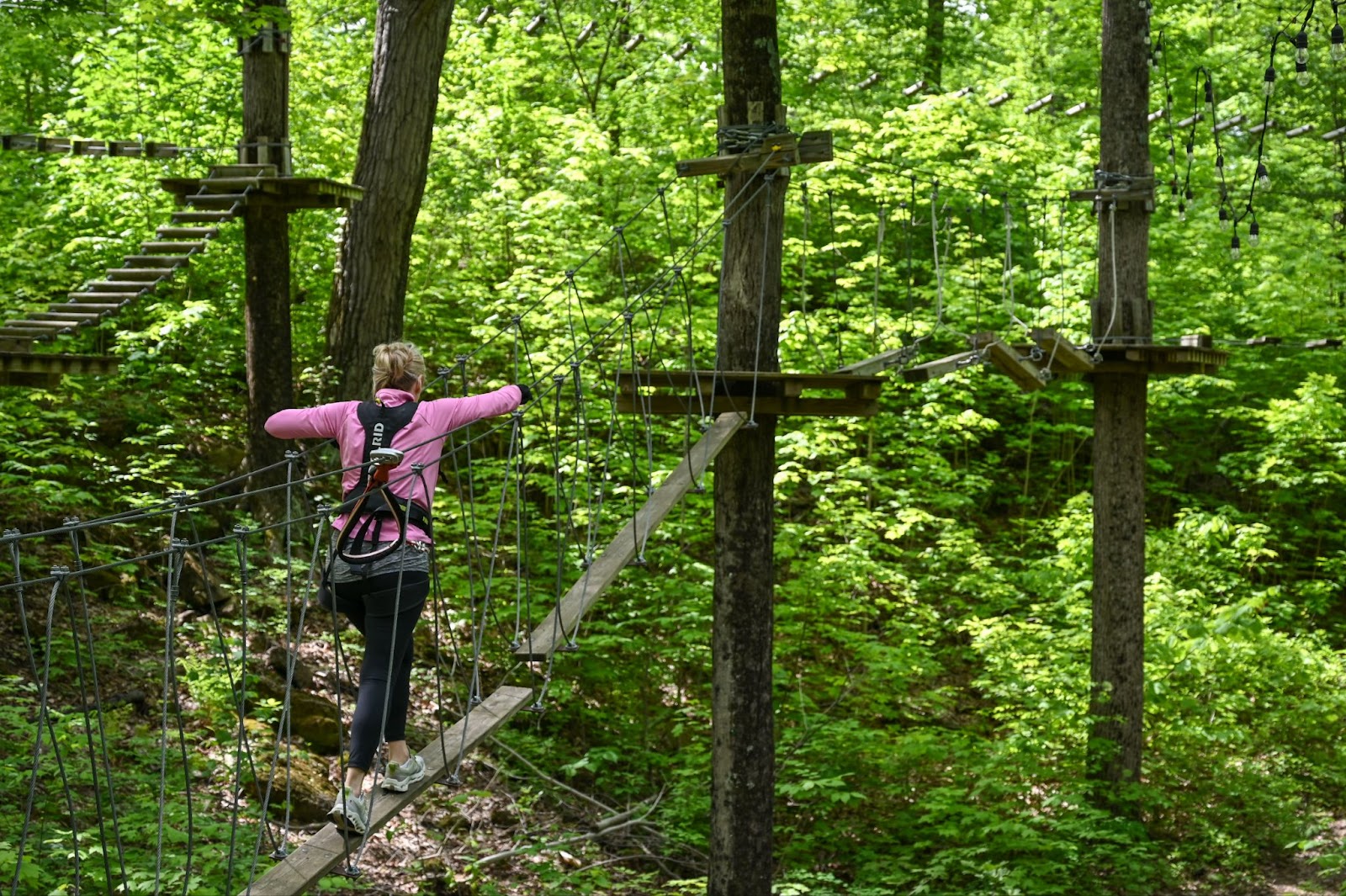 A woman photographed from behind navigating a ropes course at TreEscape Aerial Adventure Park in Vernon, NJ, carefully stepping across suspended obstacles surrounded by lush trees.