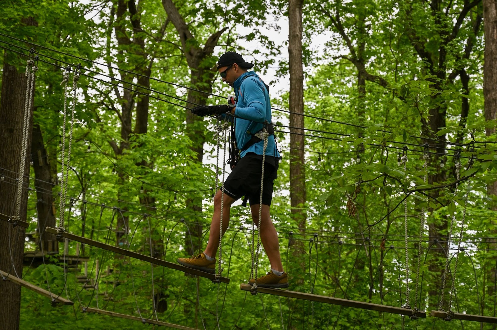 A man wearing sunglasses navigating a ropes course at TreEscape Aerial Adventure Park in Vernon, NJ, surrounded by towering trees and suspended obstacles.